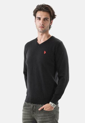 Schwarzer V-Ausschnitt-Pullover aus Strickstoff mit gerippten Bündchen und Saum; zeigt ein kleines rotes Logo auf der linken Brust; getragen mit hellgrauen Jeans.