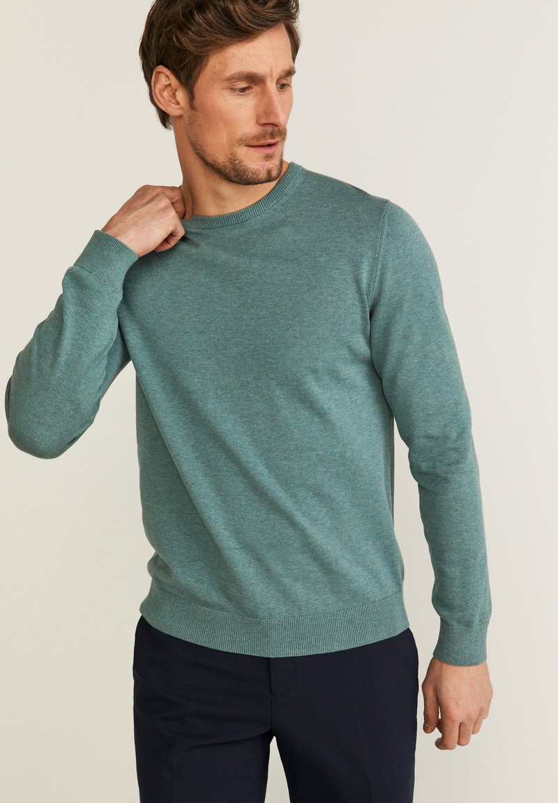 Turkos stickad tröja med rund hals, långa ärmar och ribbade muddar. Slät textur, kombinerad med mörka byxor.