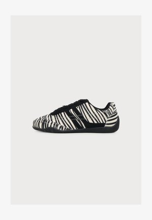 Basket basse avec rayures zèbre noires et blanches, lacets noirs et semelle noire texturée, vue de profil sur fond blanc.