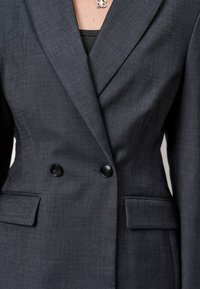 Donkergrijze blazer van textuurstof, met een dubbelgebreide ontwerp, twee knopen en zijzakken met op maat gemaakte accenten.