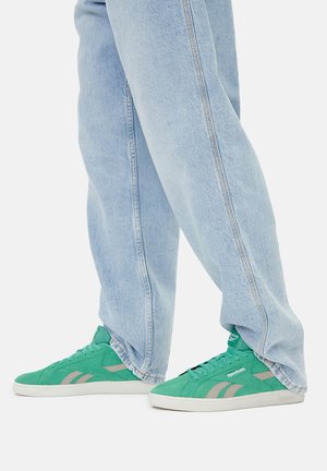 Osoba u svijetloplavim trapericama i zelenim Reebok tenisicama stoji prekriženih nogu na bijeloj pozadini.