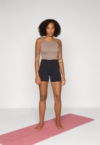 Un crop top côtelé marron et un short noir taille haute, debout sur un tapis de yoga rose. Textures lisses, design ajusté, motifs minimalistes.