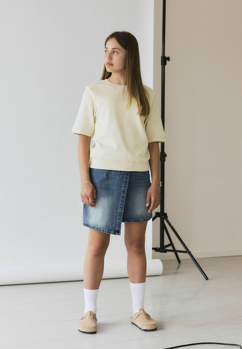 Sudadera amarilla clara con mangas cortas, combinada con una falda de mezclilla deslavada. La modelo lleva calcetas blancas y zuecos beige, de pie sobre un suelo de madera.