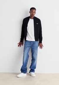 Black bomber jacket over a white t-shirt, paired with blue jeans and white sneakers. Model stands against a plain background.