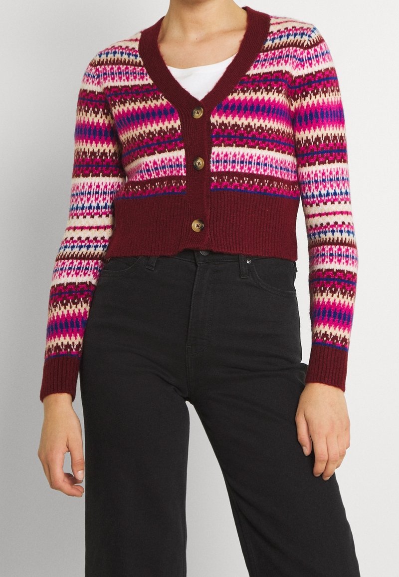 Cardigan raccourci boutonné avec des motifs géométriques rouges, roses, crème et bleus, porté sur un haut blanc et associé à un pantalon noir taille haute.