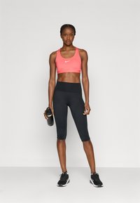Nike sportbeha in felroze met wit logo, gecombineerd met zwarte capri leggings met hoge taille. Model houdt een zwarte waterfles vast.