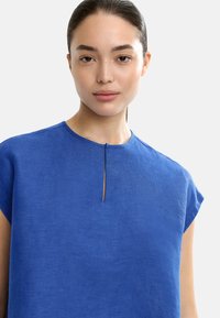 Niebieska lniana bluzka z okrągłym dekoltem i małym otwarciem z przodu. Posiada rękawy w stylu cap oraz lekko teksturowany materiał.