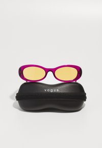 Ochelari de soare roz cu lentile galbene, având un design elegant, așezați pe o carcasă rigida neagră cu fermoar, având inscripția "vogue".