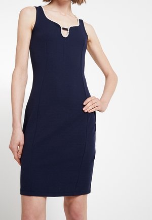 Donna che indossa un vestito aderente blu navy senza maniche con scollo quadrato rifinito con piccole perle e dettaglio di una fascia nera al centro.