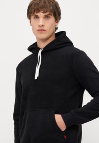 Crna flis dukserica s kapuljačom na vezanje, prednjim džepom i rebrastim manžetama. Ima mali crveni logotip na donjem rubu.