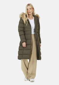 Cappotto imbottito verde oliva con cappuccio, rifinitura in pelliccia e chiusura a zip. Presenta sezioni trapuntate e un design lungo, abbinato a pantaloni beige e sneakers bianche.