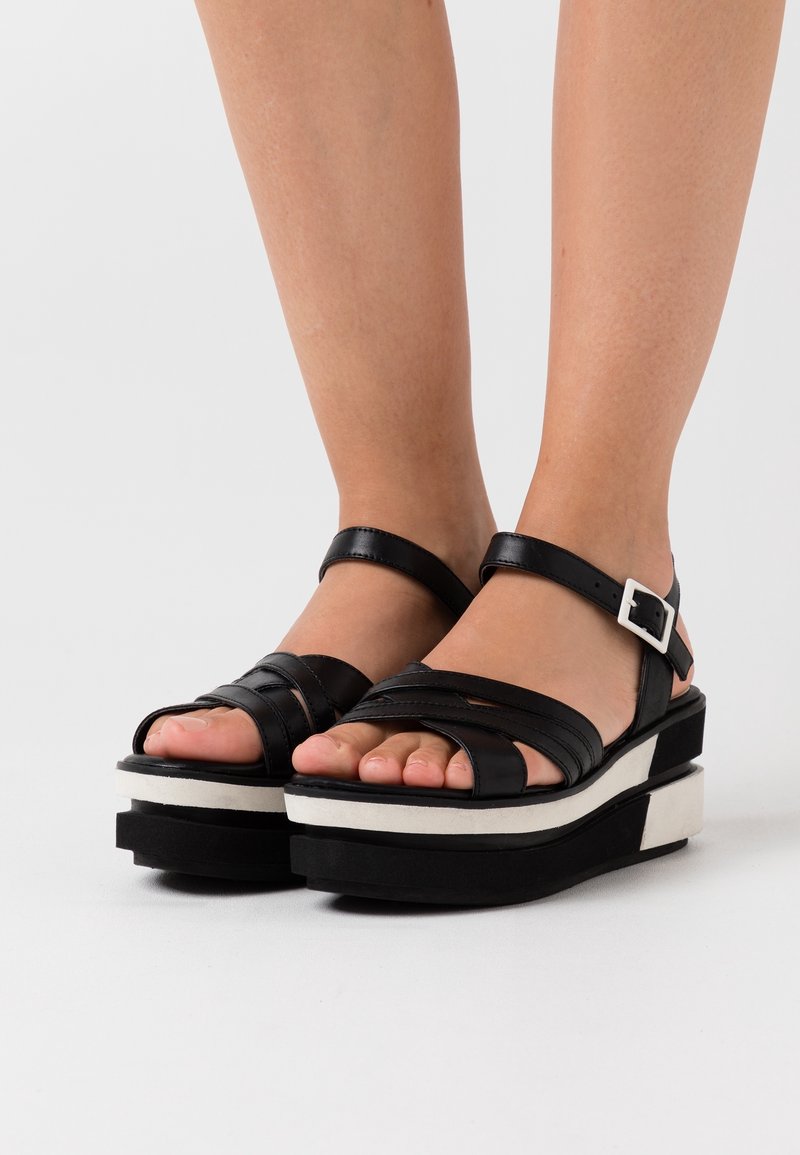 Pieds chaussés de sandales à plateforme en cuir noir avec semelles épaisses blanches et noires et brides à boucle à la cheville sur un fond uni.