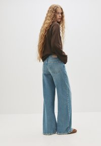 Svjetloplave široke traperice s visokim strukom, velikim stražnjim džepovima i rastrganym rubom, u kombinaciji s tamnosmeđim dugim rukavima.