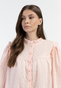 Lichtroze blouse met stippenstructuur, knoopsluiting, gerimpelde kraag en puffmouwen, gemaakt van een lichte stof.