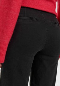 Zwarte broek van een gladde stof, met twee achterzakken en minimale stiksels, gecombineerd met een rode gebreide trui met ribbelmanchetten.