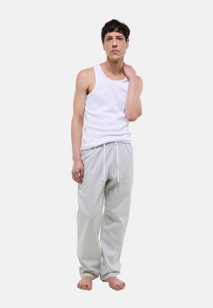 Homme debout pieds nus portant un débardeur blanc et un pantalon de pyjama gris clair rayé avec cordon de serrage, sur un fond uni.