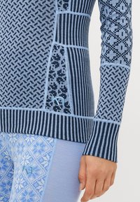 Nærbilde av en person som har på seg en blå mønstret langermet topp og leggings med varierende geometriske mønstre og synlige merkevarelogoer.