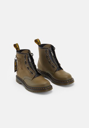 Par de botas de cuero verde oliva hasta el tobillo con ribete negro, cremallera frontal, costuras amarillas y etiquetas y tiradores de marca sobre un fondo blanco.