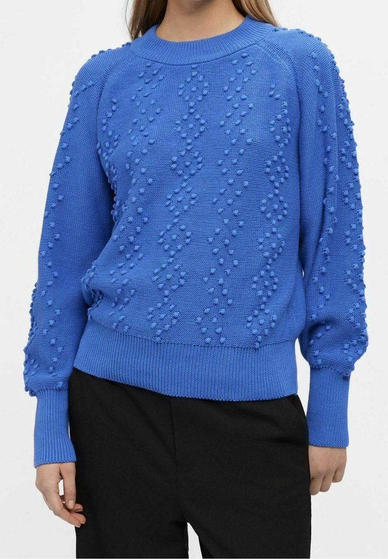 Femme portant un pull en tricot bleu texturé avec des motifs de points en relief et un pantalon noir, debout devant un fond uni.