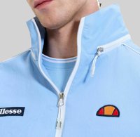 Młody mężczyzna ubrany w jasnoniebieską kurtkę z zamkiem błyskawicznym oraz białym zamkiem, z naszywkami logo ellesse na piersi.