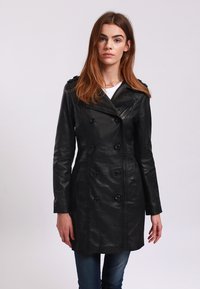 Cappotto trench in pelle nera con bottoni doppi, vestibilità sartoriale e colletto affilato. Tessuto morbido con dettagli di cucitura ben visibili.