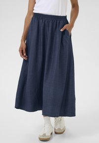 Fustă maxi de culoare bleumarin, dintr-un material texturat, cu talie elastică și buzunare laterale, asortată cu adidași albi și șosete până la glezne.