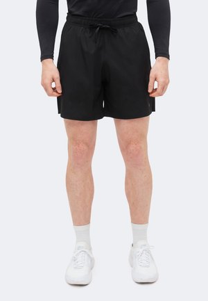 Mand iført sorte atletikshorts, hvide sokker og hvide løbesko, der står mod en ensfarvet baggrund.