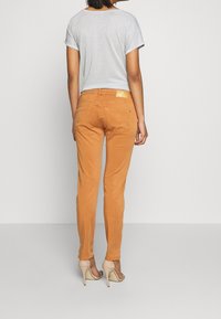 Pantalon slim en velours côtelé orange avec une texture subtile, doté de poches arrière et d'une étiquette décorative à la taille ; porté avec un haut gris clair.