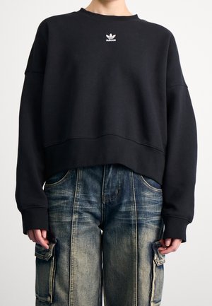 Osoba ubrana w czarną skróconą bluzę Adidas oraz wyblakłe niebieskie dżinsy typu cargo z kieszeniami na klapki, stojąca na neutralnym tle.