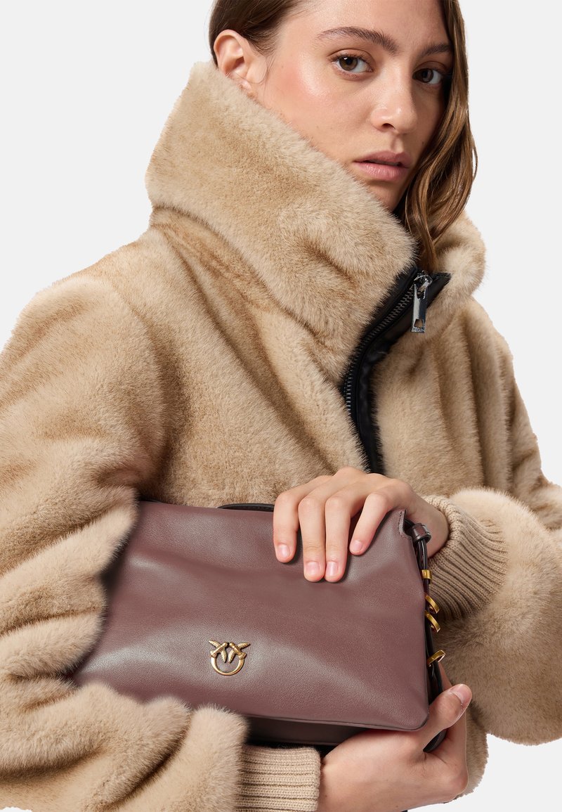 Borsa in pelle marrone con logo dorato, tenuta da una persona che indossa una giacca beige e pelosa con colletto alto e zip nera.