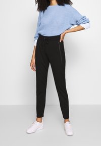 Svarta joggerbyxor med snörning i midjan och sidofickor med dragkedja, i kombination med en ljusblå stickad tröja och vita sneakers.