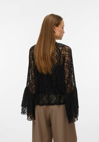 Svart blondetopp med lange klokkeermer, gjennomskinnelig tekstur, blomsterdekor, lagdelt design, brukt over beige bukser. Synlig detalj bak.