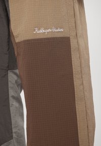 Παντελόνια με μπλοκ χρωμάτων, ελαφριά και υφασμένα με υφή σε μπεζ, καφέ και γκρι. Διαθέτουν λευκή κεντητή επιγραφή "KidSuper Studios" στο πάνω μέρος του ποδιού.