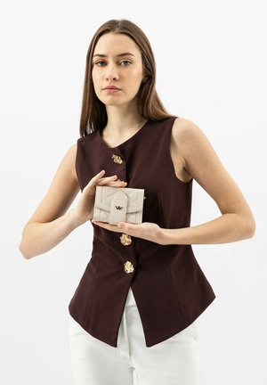 Beige leren portemonnee met een gequilt patroon en gouden accenten, vastgehouden door een model in een donkerpaarse mouwloze top tegen een witte achtergrond.