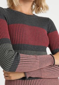 Femme portant un pull en tricot côtelé avec des rayures horizontales en gris foncé, bordeaux, vert olive et rose poussiéreux, les bras croisés.
