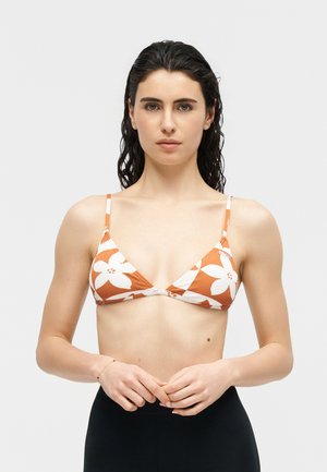 Femeie cu păr creț și ud, de culoare închisă, purtând un top de bikini triunghiular portocaliu și alb cu motive florale, stând pe un fundal simplu, deschis.