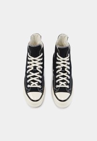 Paire de baskets montantes en toile noire avec lacets blancs et embouts en caoutchouc, vues de dessus sur un fond clair.