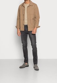 Beige bomuldsjakke med brystlommer, båret over en lys beige skjorte og mørkegrå jeans; grå slip-on sko med minimal detaljering.
