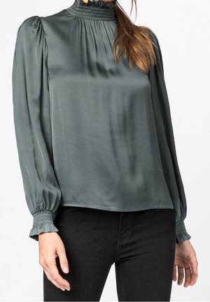 Femme portant un chemisier en satin vert foncé à manches longues avec col haut et poignets smockés, associé à un pantalon noir ajusté.