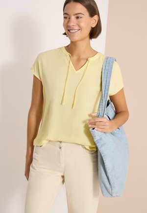 Uśmiechnięta kobieta w bladożółtej koszulce z krótkim rękawem i jasnobeżowych spodniach niesie dużą jasnoniebieską dżinsową torbę na ramię.