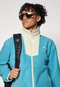 Veste en polaire bleu clair avec des accents crème, col haut, fermeture éclair devant et poches latérales. Le modèle porte des lunettes de soleil marron et un sac à dos noir.