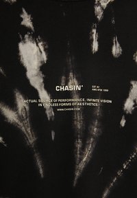 Tissu noir avec motifs tie-dye blancs entourant un texte centré : « CHASIN', EST. 92, AMS-AFW 1059, source réelle de performance, vision infinie.