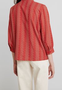 Rød mønstret bluse med et fiskebensdesign, der har trekvartlange ærmer, sammensnørede manchetter og en høj halsudskæring. Stoffet virker let.