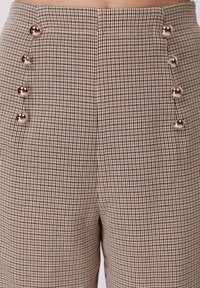 Καρό παντελόνι με ψηλή μέση σε αποχρώσεις καφέ και κρεμ, με γυαλιστερά ασημένια κουμπιά στην αριστερή πλευρά και λείο ύφασμα.
