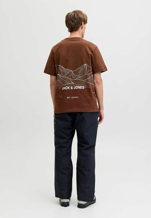T-shirt marron à manches courtes avec un graphique de ligne de montagne blanche et du texte. Associé avec un pantalon noir. Fond uni, mise au point sur les détails du vêtement.