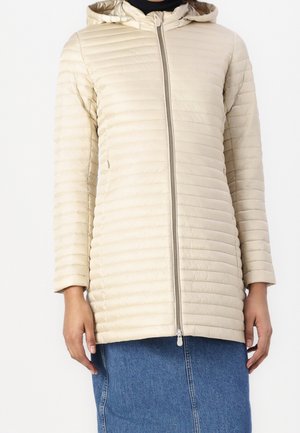 Veste matelassée beige avec capuche et fermeture éclair devant, portée avec une jupe en jean bleue, montrée des épaules aux hanches.
