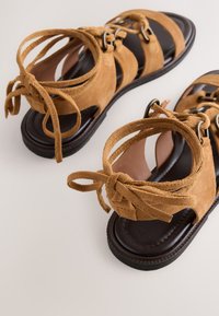 Sandales en cuir suédé marron avec semelles noires, comportant plusieurs lanières et lacets autour de la cheville sur fond blanc.