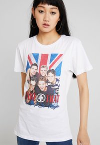 Junge Frau mit langen schwarzen Haaren, die große Creolen trägt und ein weißes T-Shirt mit einem Foto einer Boyband und einem Union-Jack-Hintergrund anhat.