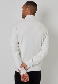 Weißer Pullover mit hohem Kragen, gerippten Bündchen und Saum. Tragen mit hinter dem Rücken verschränkten Händen. Glatte Textur, taillierte Passform.