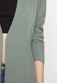 Ljusgrön långärmad jacka med rynkningar vid ärmsluten, med en djup V-ringning och textil material, som bärs över en svart topp.